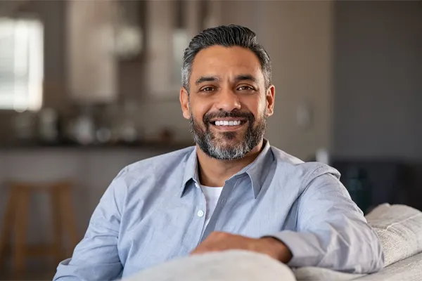 BioTE-Testosterone-Pellet-Clinic A mature South Asian man seated comfortably on a couch, smiling and looking directly at the camera satisfied with BioTE Testosterone Pellet Therapy offered by Jennifer Jose-Cappola, MD in Stamford.
