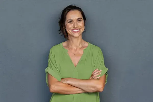 Hormones-for-Women-Doctor A woman in a green shirt stands smiling against a gray-blue wall, pleased with her hormone therapy from Jennifer Jose-Cappola, MD in Stamford.