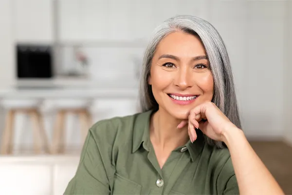 Menopause-Clinic An asian woman with long gray hair and a green shirt sitting and smiling on her couch, happy with her menopause care from [NAME} in Stamford.